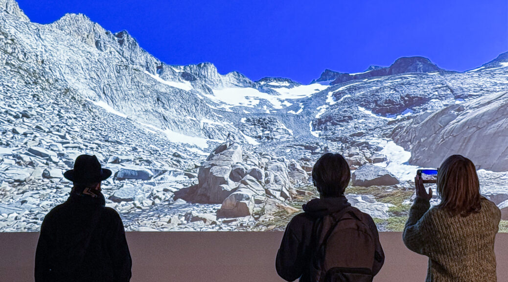 Three people at an art exhibit stand with backs to camera, regarding a projected image of high mountain melting glaciers.