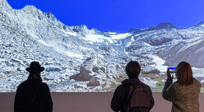 Three people at an art exhibit stand with backs to camera, regarding a projected image of high mountain melting glaciers.